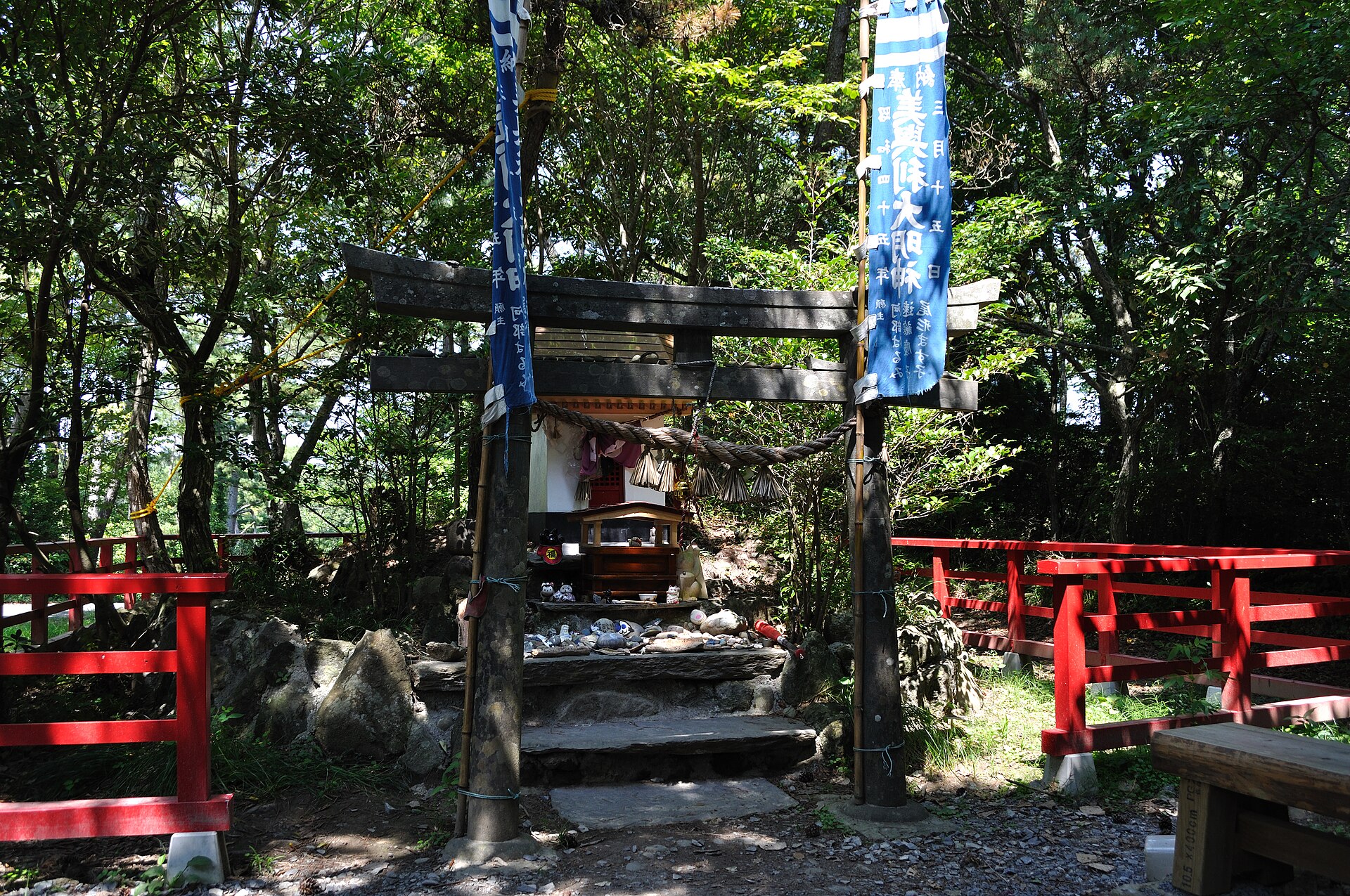 A stone torii with blue banners and a small shrine behind it, its steps crowded with maneki-neko figures and offerings